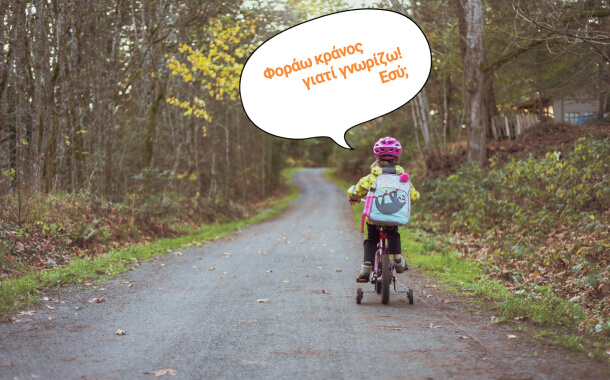 Κοριτσάκι κάνει ποδήλατο σε χωματόδρομο με δέντρα φορώντας κράνος ασφλείας. Πάνω από το κεφάλι της υπάρχει ένα συννεφάκι με το μήνυμα: «Φοράω κράνος γιατί γνωρίζω! Εσύ;»